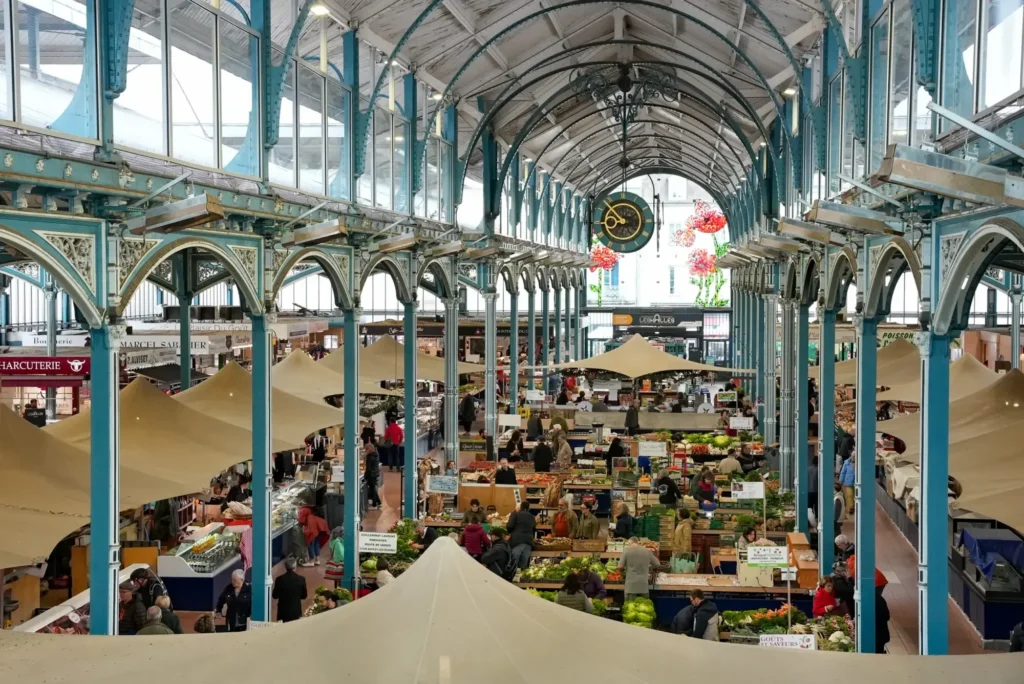 Vue plongeante d'un marché couvert historique animé, avec piliers métalliques bleus et nombreux étals.