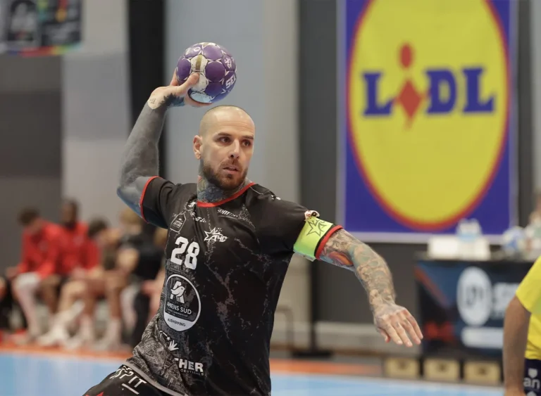 Joueur de handball tatoué en maillot noir armant son tir avec un ballon violet.