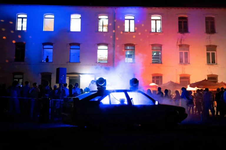 Silhouette d'une voiture avec fumée et projecteurs bleus devant un bâtiment éclairé de couleurs vives.