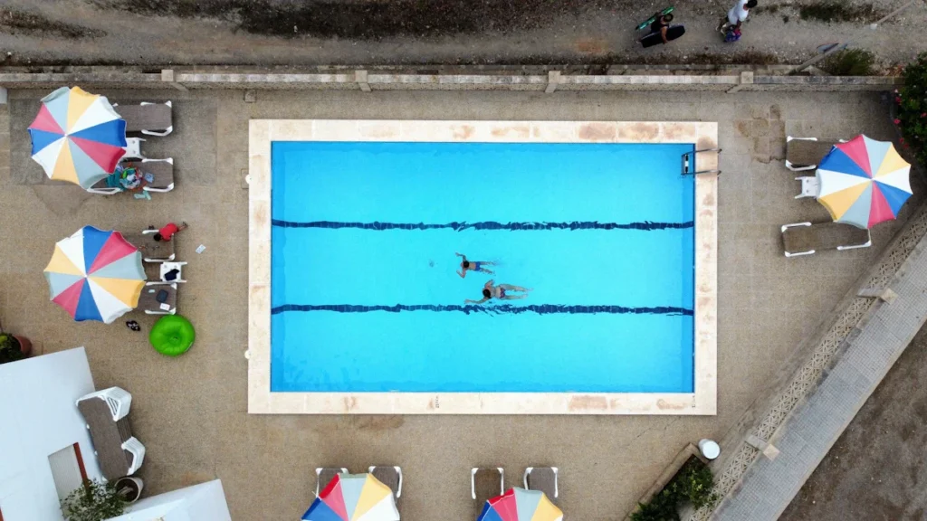 Vue de dessus d'une piscine avec des couloirs de nage, entourée d'un solarium aménagé avec des transats et des parasols colorés.