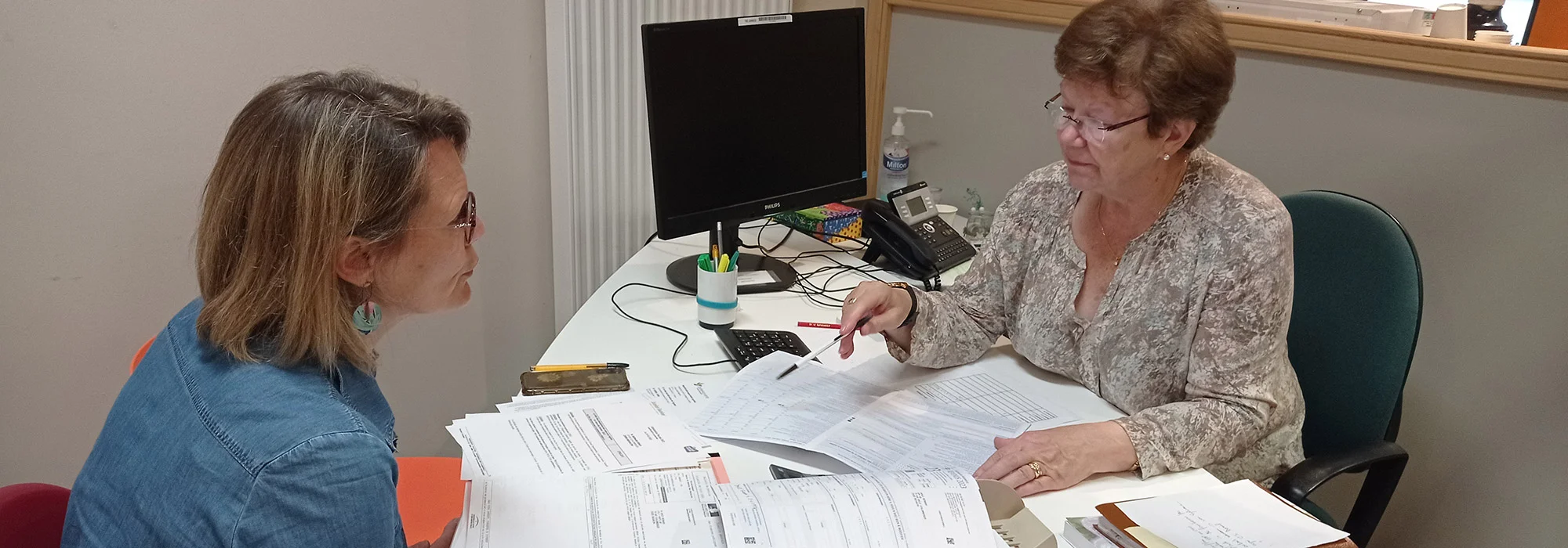Une femme à lunettes pointe un document pour une cliente dans un bureau administratif lumineux.