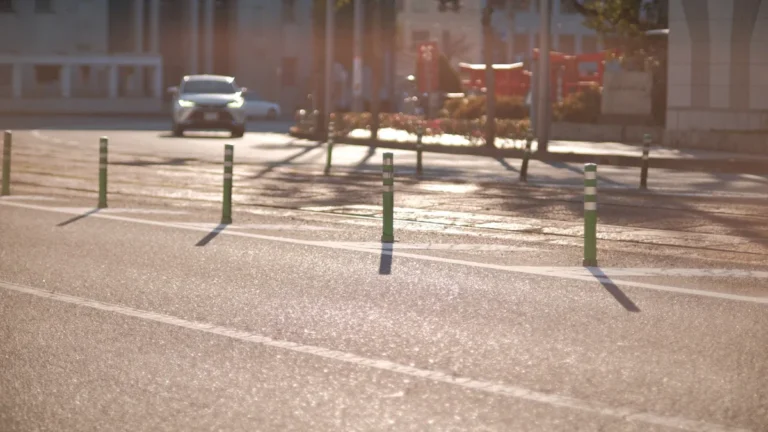 Une voiture blanche circulant sur une voie urbaine bordée de poteaux de signalisation sous une lumière matinale.