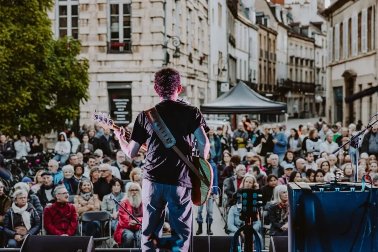 Musicien de dos jouant de la guitare devant une foule assise dans une rue ancienne.