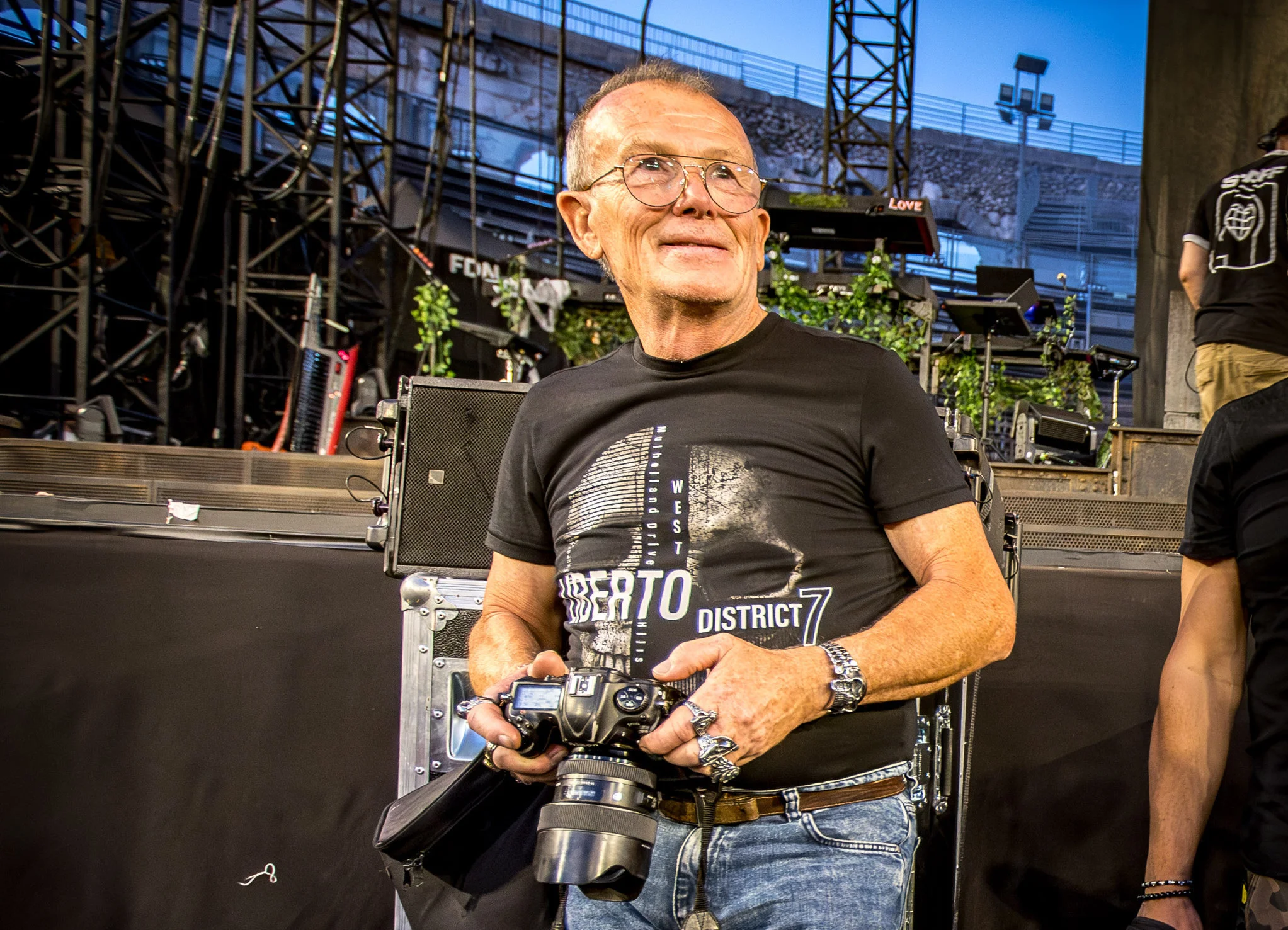 Homme souriant à lunettes tenant un appareil photo reflex devant une scène de concert en extérieur.