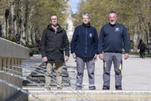 Trois hommes posent derrière une fontaine d'eau dans une allée bordée d'arbres ensoleillée.