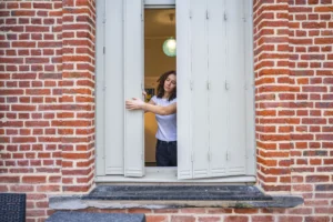 Femme aux cheveux bouclés ouvrant des volets gris sur une façade de briques rouges.