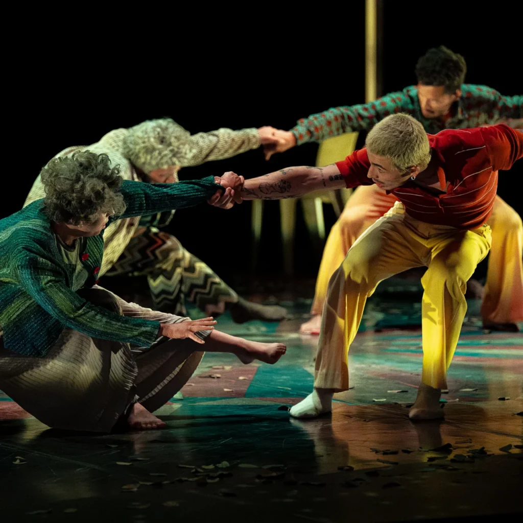 Des performeurs en vêtements colorés s'agrippent les mains lors d'une chorégraphie dynamique sur scène sombre.