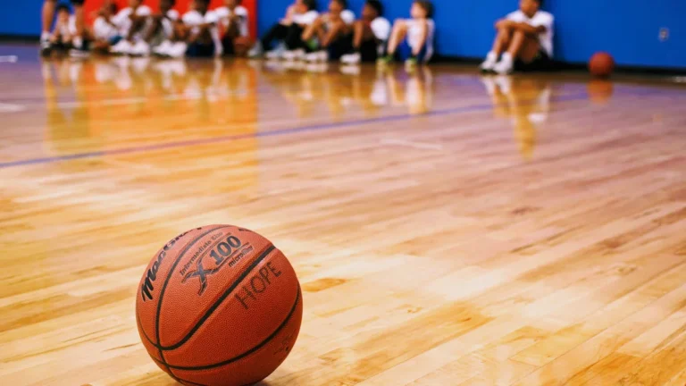Un ballon de basket posé sur le parquet d'un gymnase avec des enfants assis en arrière-plan flou.