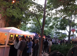 Foule réunie en extérieur sous des arbres et une tente blanche ornée de lampions colorés.