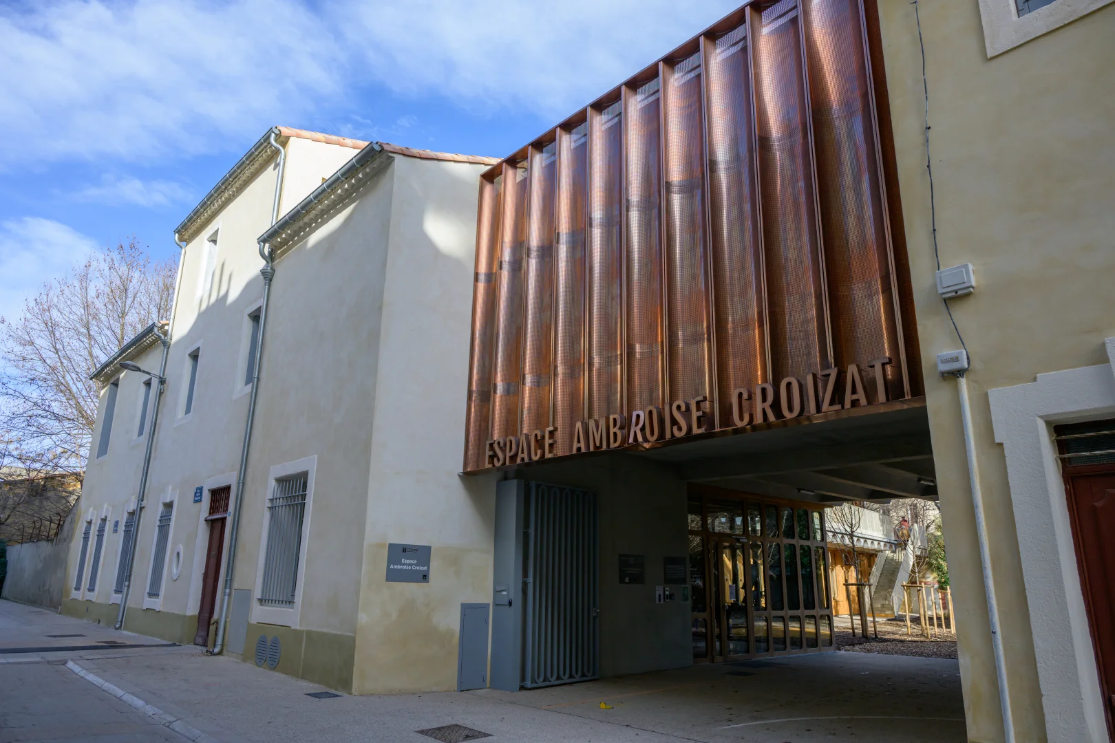 Façade d'un bâtiment beige avec une structure en métal cuivré marquée Espace Ambroise Croizat.