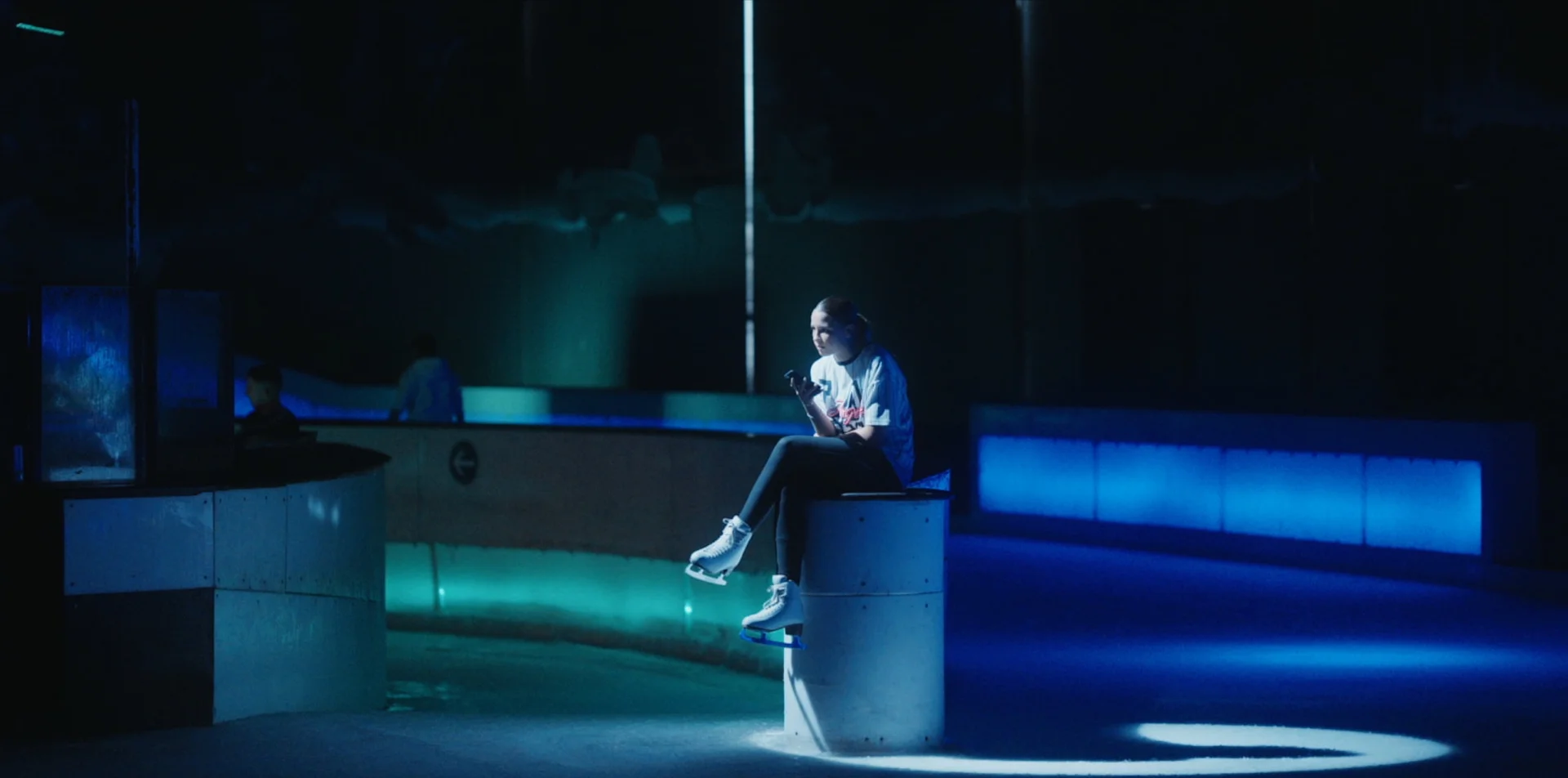 Patineuse assise sur un pilier blanc regarde son smartphone dans une patinoire sombre et bleue.