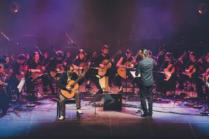 Un orchestre de guitares acoustiques jouant sur scène sous un éclairage tamisé violet et rose.