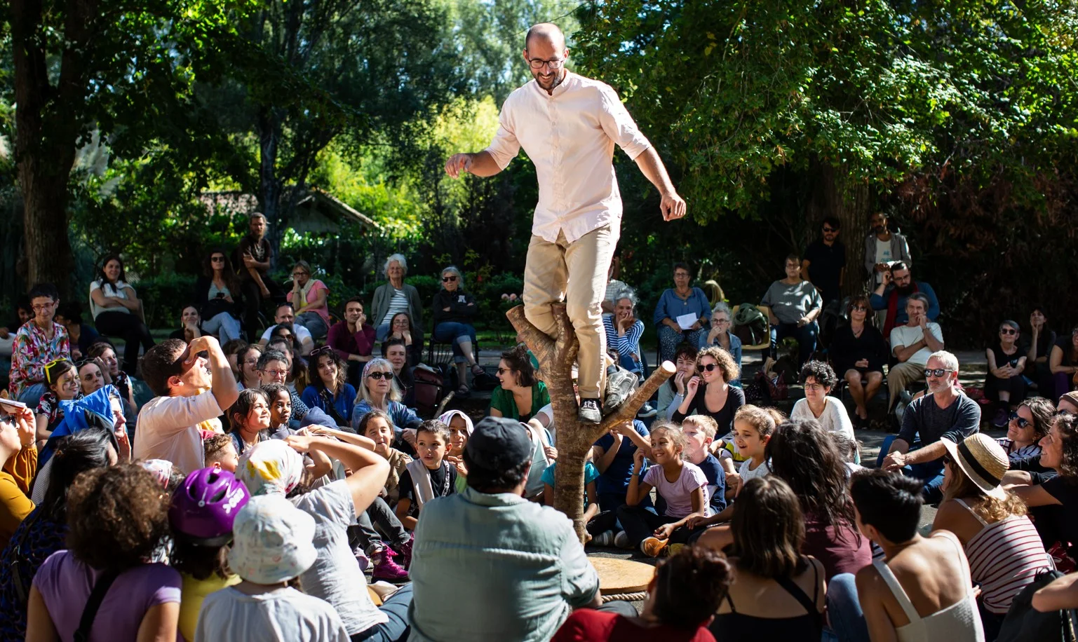 Un acrobate en équilibre sur une branche devant une foule attentive dans un parc ensoleillé.