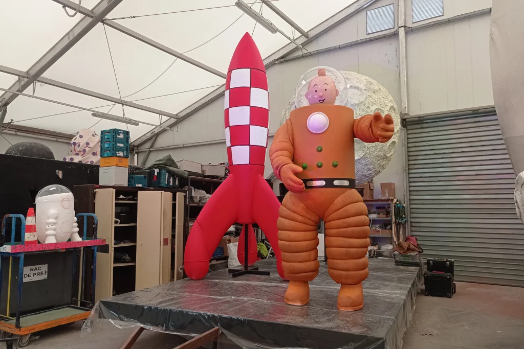 Grande statue de Tintin en scaphandre orange devant sa fusée rouge et blanche dans un entrepôt.