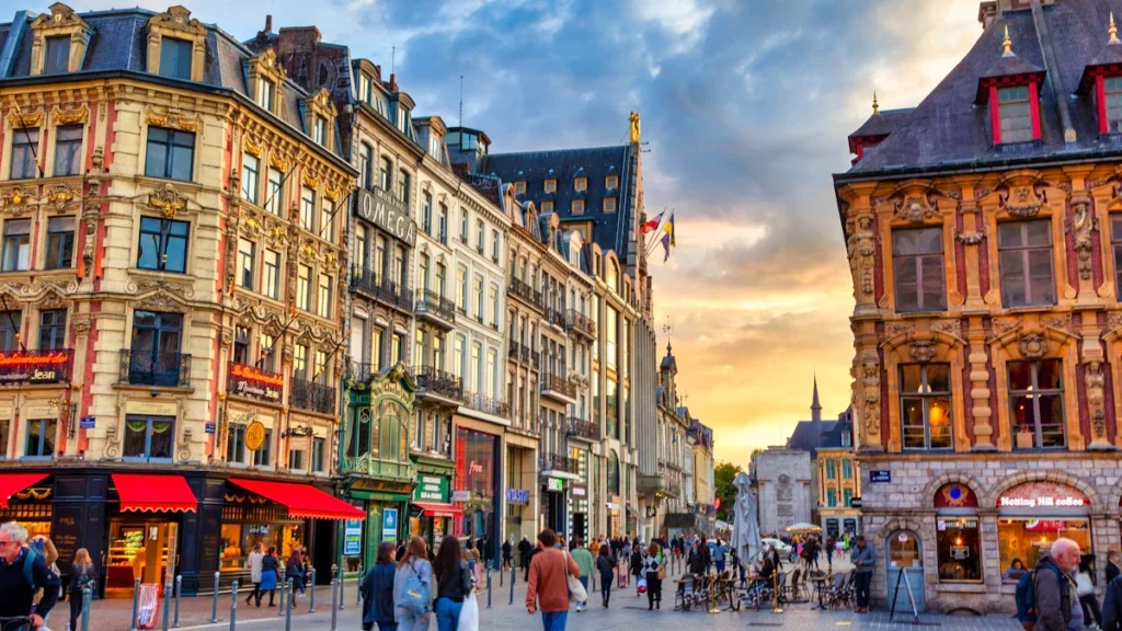 Des passants et des terrasses de cafés dans une rue commerçante du centre-ville de Lille sous un ciel de fin de journée.