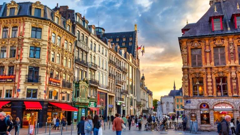 Des passants et des terrasses de cafés dans une rue commerçante du centre-ville de Lille sous un ciel de fin de journée.