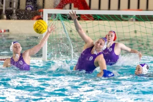 Joueuses de water-polo en maillot violet défendant leur but contre un ballon jaune.