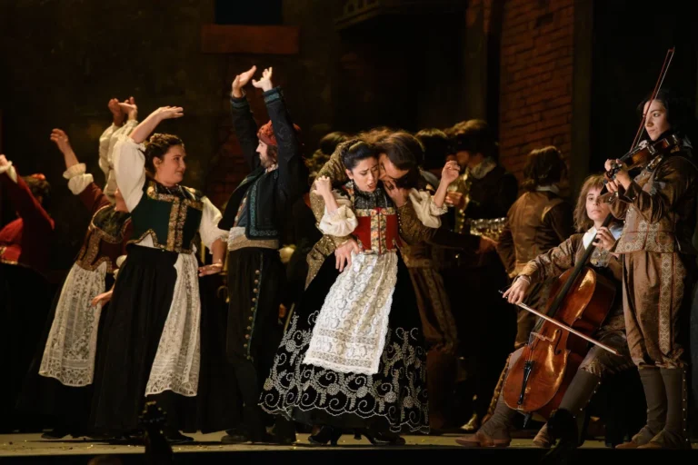 Acteurs d'opéra en costumes d'époque sur scène avec des danseurs et des musiciens d'orchestre.