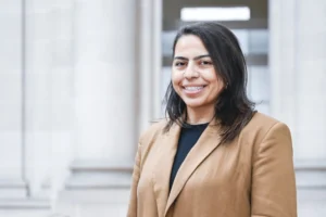 Une femme souriante aux cheveux noirs portant un blazer beige devant des colonnes en pierre.