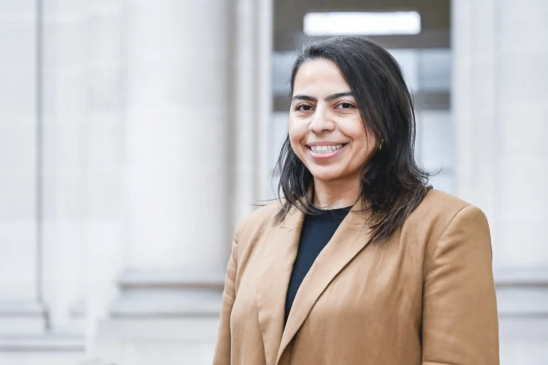 Une femme souriante aux cheveux noirs portant un blazer beige devant des colonnes en pierre.