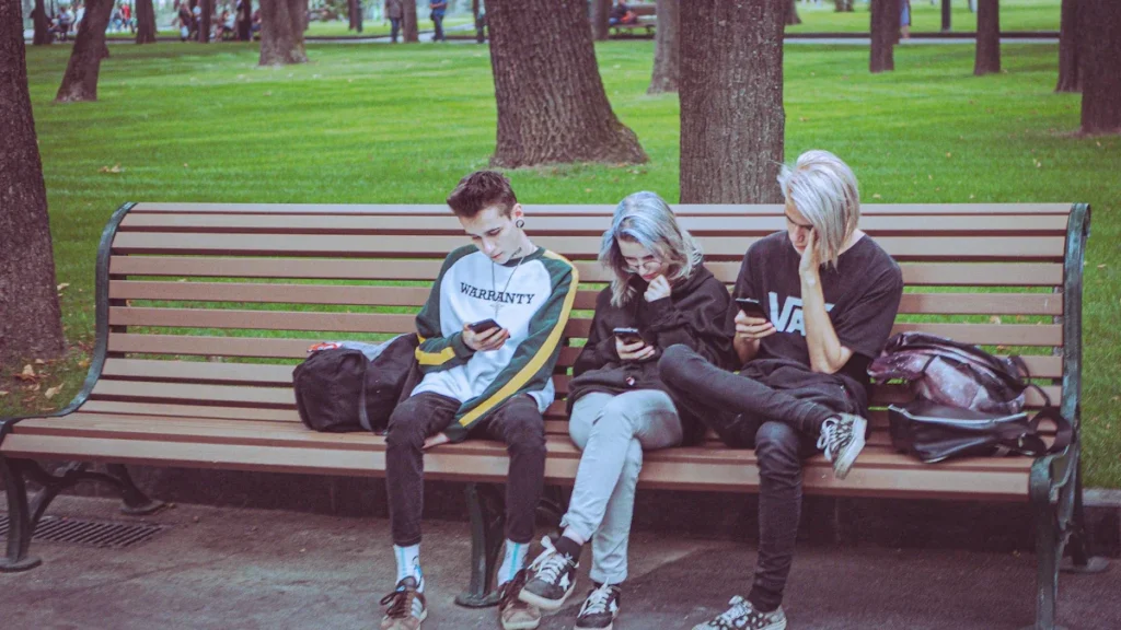 Trois adolescents assis sur un banc de bois dans un parc, occupés avec leurs téléphones portables.