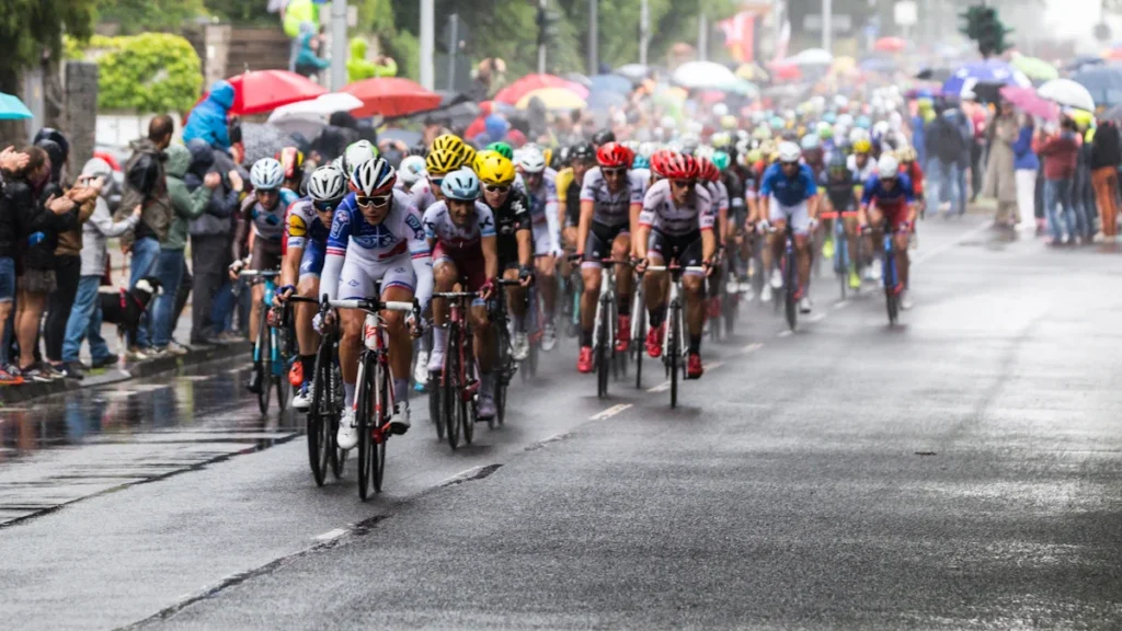Peloton de coureurs cyclistes sous la pluie lors d'une compétition urbaine bordée de spectateurs avec parapluies.