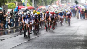 Peloton de coureurs cyclistes sous la pluie lors d'une compétition urbaine bordée de spectateurs avec parapluies.