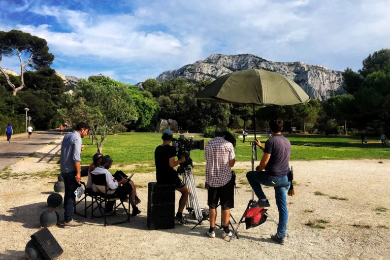 Équipe de tournage avec caméra professionnelle et grand parasol devant une montagne rocheuse imposante.