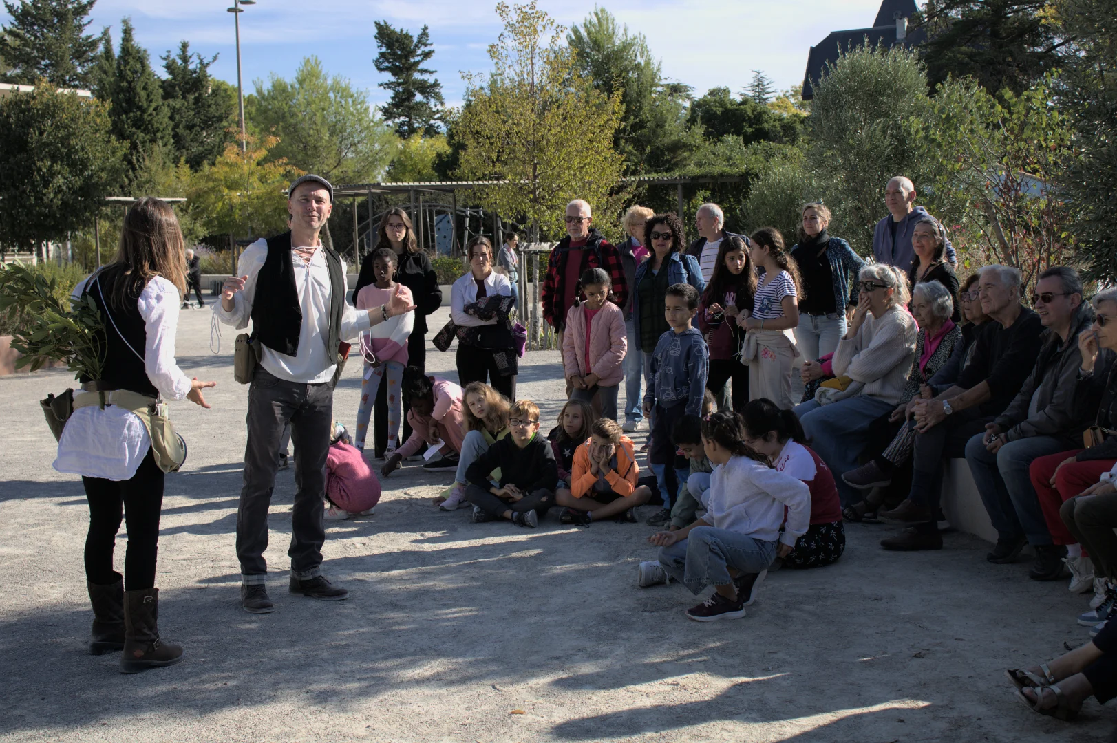 Deux comédiens en costume s'adressent à un public d'enfants et d'adultes dans un parc ensoleillé.