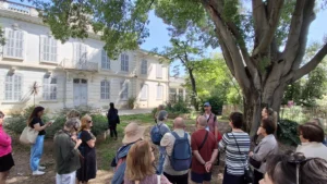 Groupe de personnes écoutant un guide devant un grand bâtiment blanc aux volets gris.