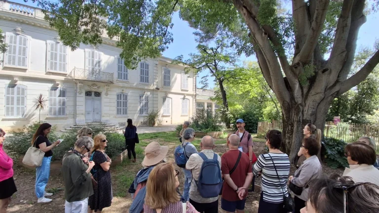 Groupe de personnes écoutant un guide devant un grand bâtiment blanc aux volets gris.