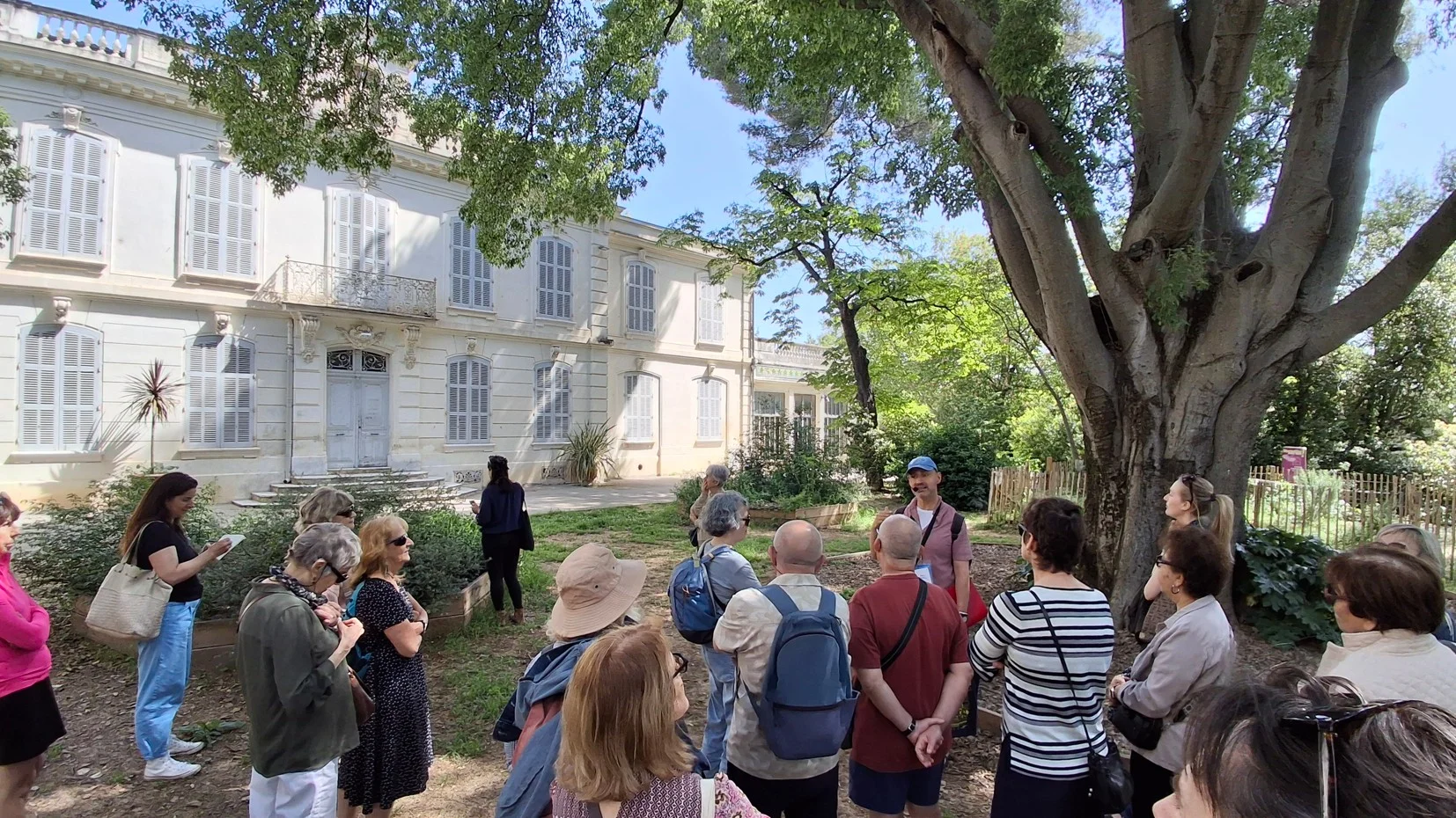 Groupe de personnes écoutant un guide devant un grand bâtiment blanc aux volets gris.