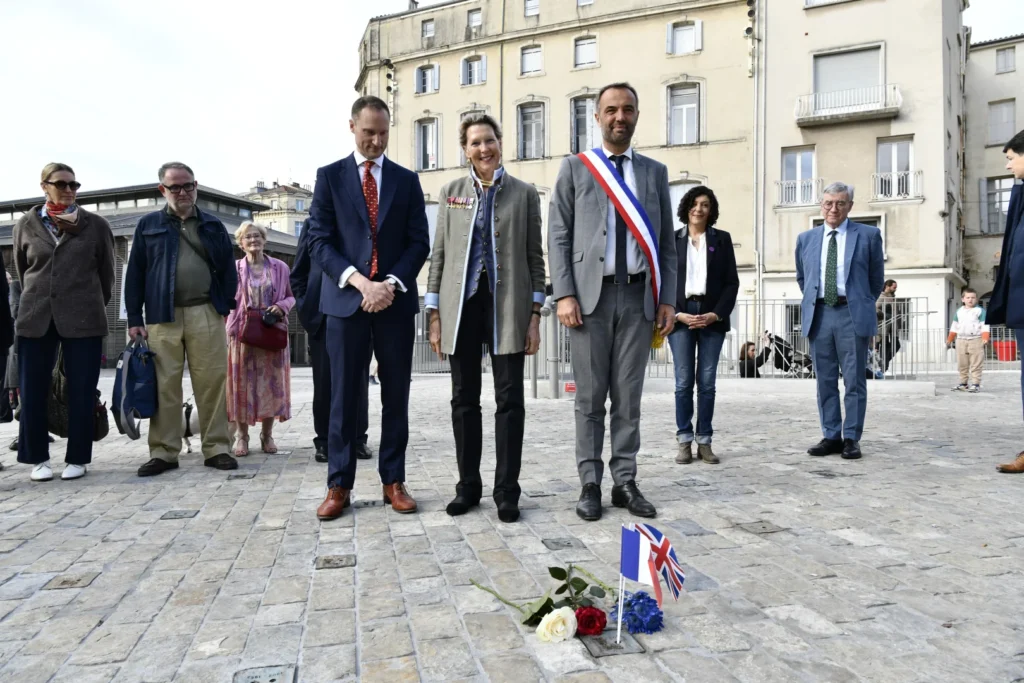 Des officiels debout sur une place pavée devant des roses et des petits drapeaux au sol.