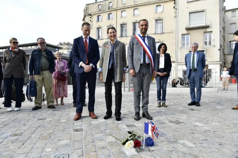 Des officiels debout sur une place pavée devant des roses et des petits drapeaux au sol.