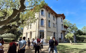 Groupe de personnes devant une grande villa en pierre entourée d'arbres sous un soleil éclatant.