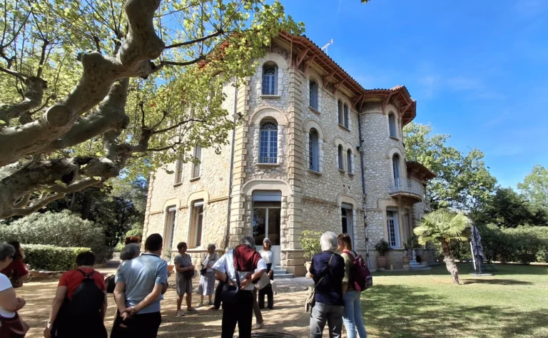 Groupe de personnes devant une grande villa en pierre entourée d'arbres sous un soleil éclatant.