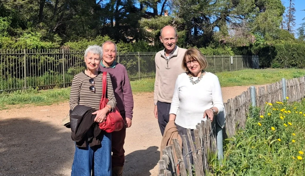 Quatre adultes sourient sur un sentier ensoleillé près d'un jardin fleuri et d'une clôture en bois.