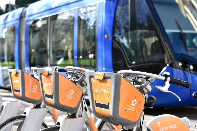 Vélos orange Vélomagg alignés au premier plan avec un tramway bleu flou en arrière-plan.