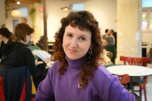 Portrait d'une femme souriante aux cheveux bouclés roux portant un pull à col roulé violet.