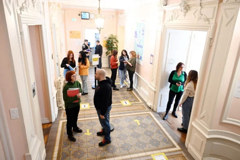Plusieurs personnes discutent dans un couloir décoré de carrelage ancien et de moulures blanches.