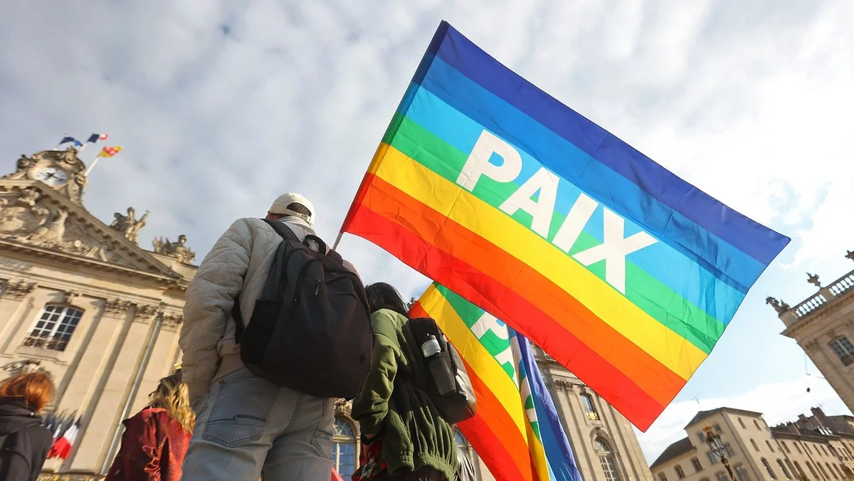 Un homme de dos brandissant un drapeau arc-en-ciel marqué 'PAIX' devant un bâtiment classique français.