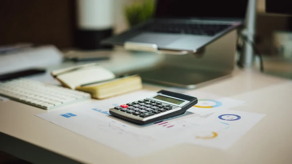Une calculatrice posée sur des graphiques financiers et des factures à côté d'un ordinateur portable dans un bureau.