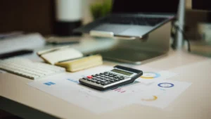Une calculatrice posée sur des graphiques financiers et des factures à côté d'un ordinateur portable dans un bureau.