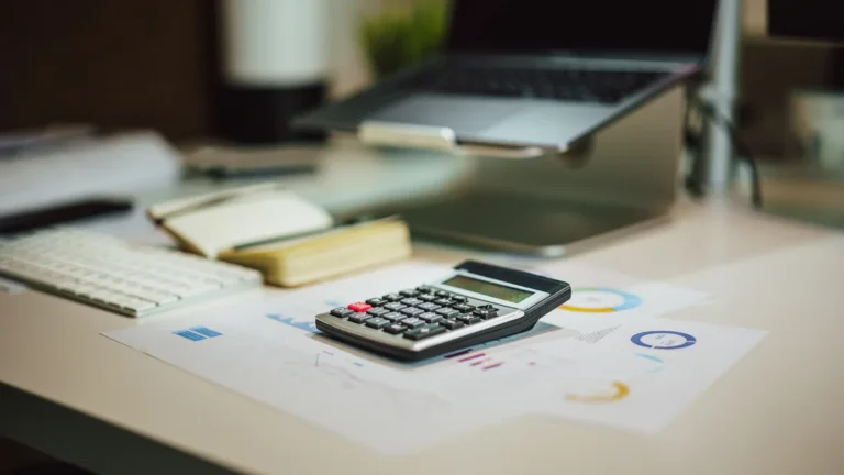 Une calculatrice posée sur des graphiques financiers et des factures à côté d'un ordinateur portable dans un bureau.