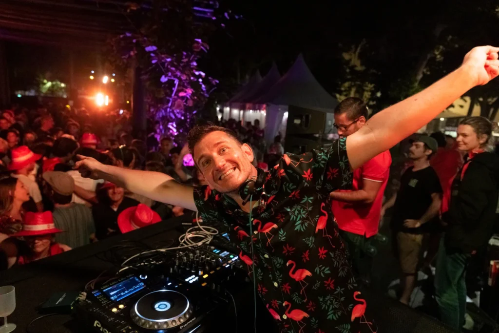 Un DJ souriant en chemise à motifs flamants roses lève les bras devant ses platines.