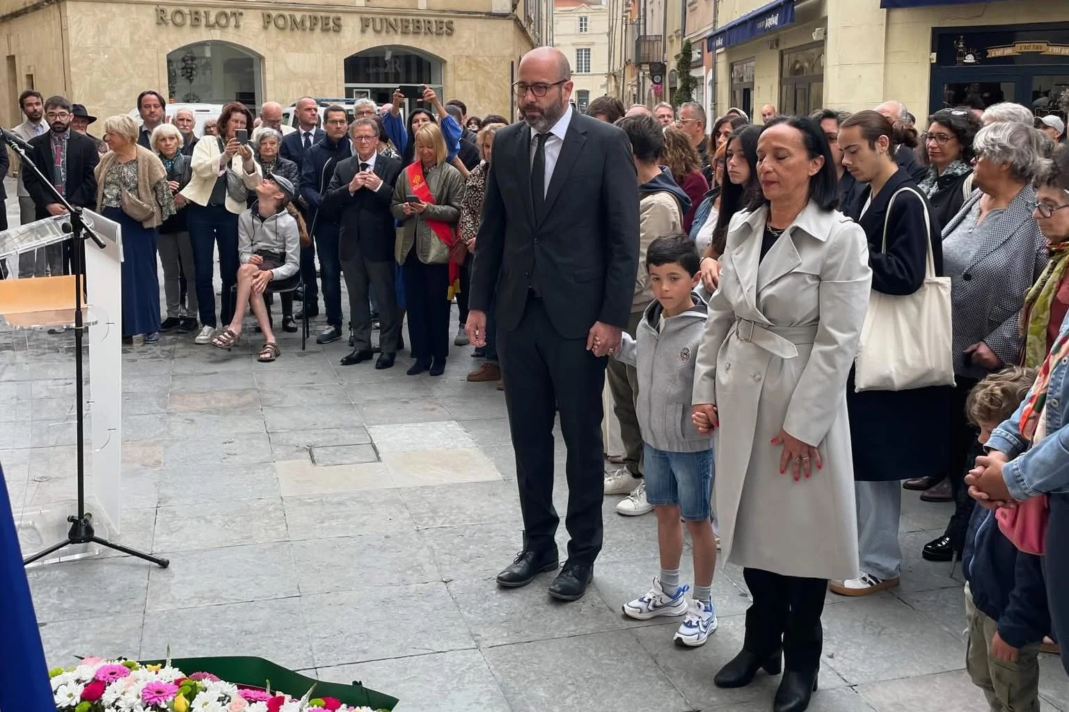 Un homme, un enfant et une femme debout solennellement devant une foule et des fleurs.