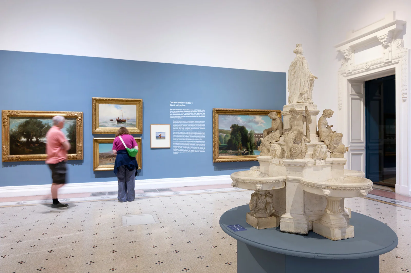 Galerie d'art avec des tableaux de paysages et une grande sculpture fontaine au premier plan.
