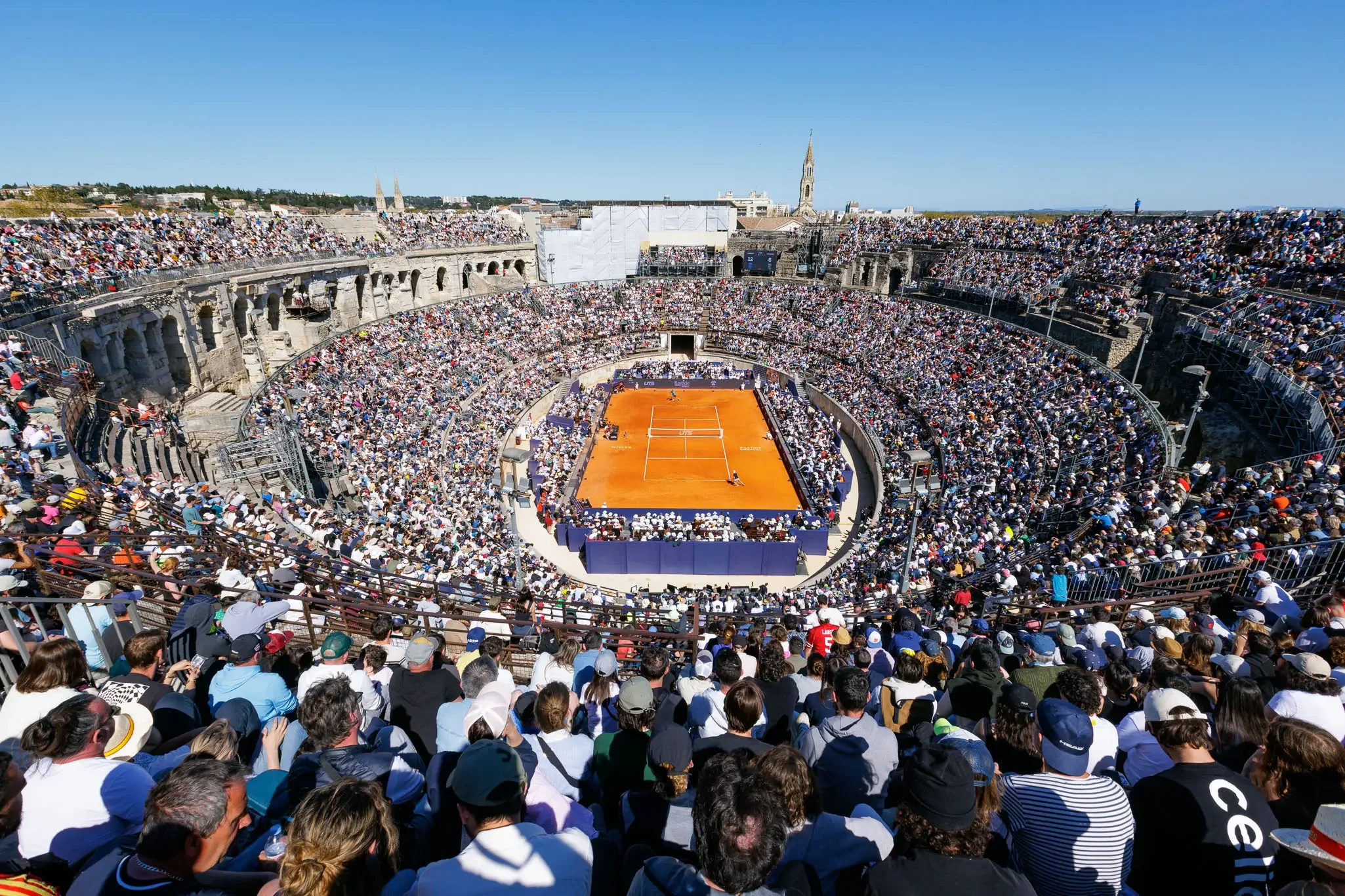 Vue aérienne d'un court de tennis en terre battue dans un amphithéâtre romain bondé de spectateurs.