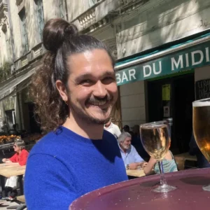 Homme souriant avec un chignon et pull bleu devant la terrasse du Bar du Midi.