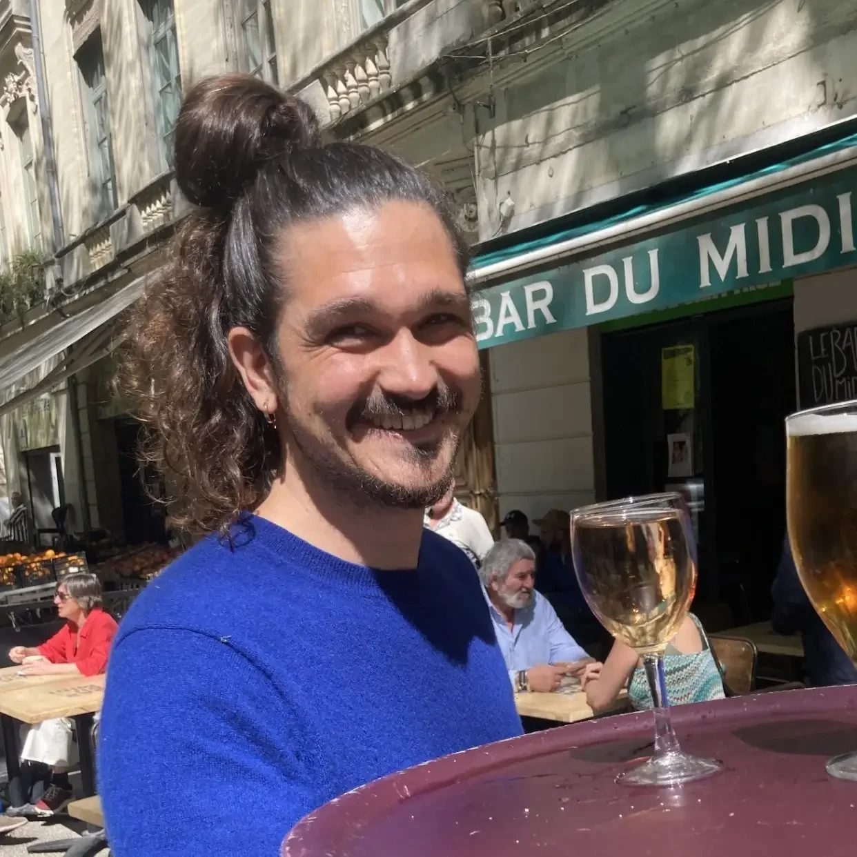 Homme souriant avec un chignon et pull bleu devant la terrasse du Bar du Midi.